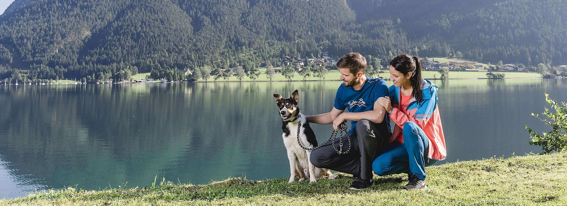 Ein Mann und eine Frau sitzen mit einem Hund am Ufer des Achensee.